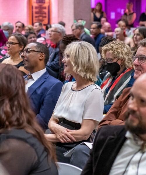 A diverse group of men and women sitting in rows at an indoor event. Some are smiling or engaged in conversation. The background is warmly lit with purple and orange lights, adding a vibrant ambiance to the scene.
