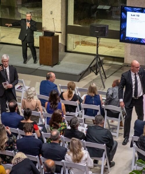 An auction in progress at a gallery, with an auctioneer standing behind a podium and gesturing toward a displayed painting. Attendees are seated in white chairs, some observing, others engaging with raised hands, while two men stand nearby, helping facilitate the auction.
