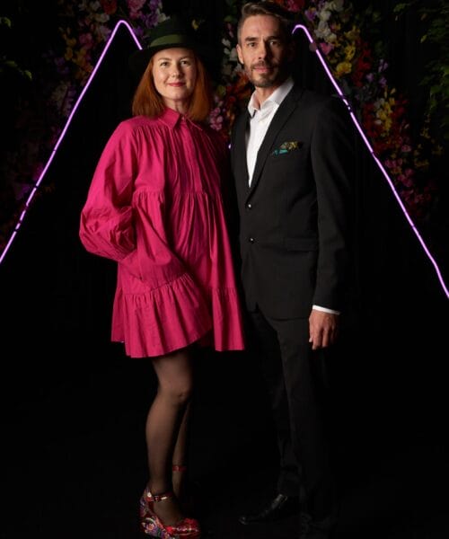 A woman in a bright pink dress, floral platform shoes, and a black hat stands next to a man in a black suit with an open white shirt. They stand against a backdrop of colorful flowers and neon lights in a dark setting, smiling at the camera.