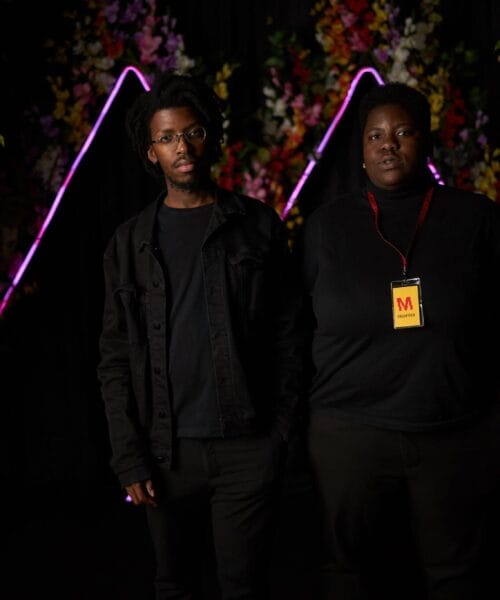 Two individuals stand side by side against a dark backdrop adorned with colorful floral decorations and neon geometric designs. Both are dressed in black; the person on the right wears a lanyard with a visible ID badge. The atmosphere appears creative and artistic.