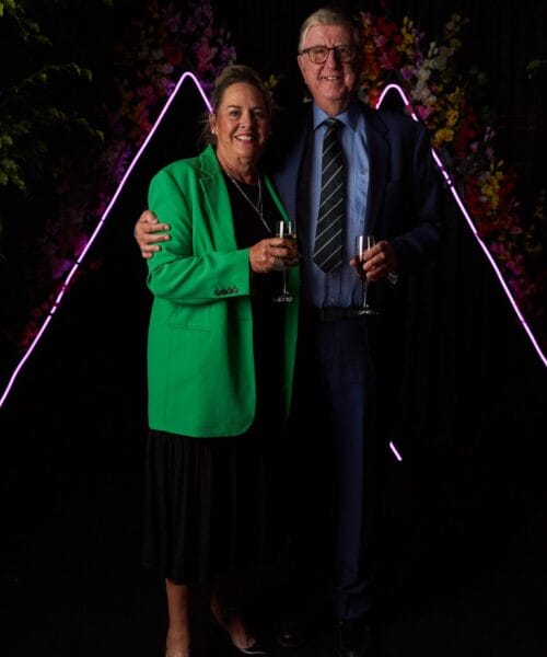 A smiling couple stands close together, holding glasses of wine. The woman wears a green blazer over a black dress, while the man wears a blue suit and striped tie. They are against a dark backdrop with pink neon triangle lights and colorful floral decorations.