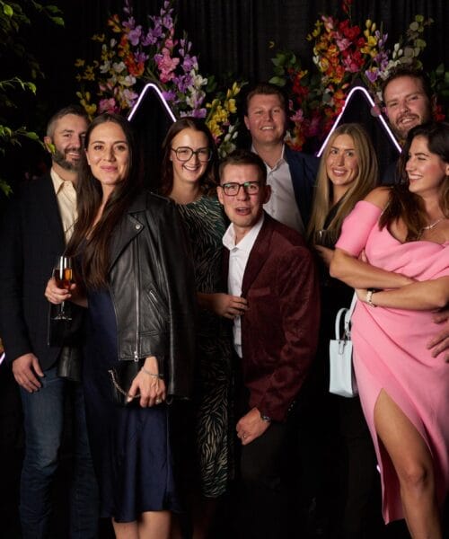 A group of eight people pose together at an event, standing in front of a dark backdrop adorned with colorful flowers and neon lights shaped like peaks. The individuals are dressed in semi-formal attire, with some holding drinks and smiling at the camera.