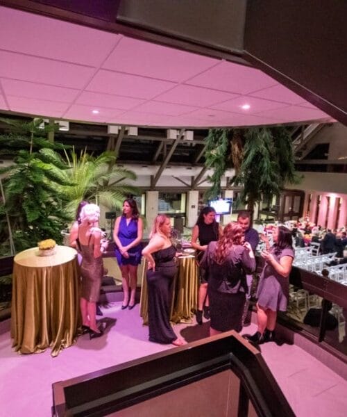 A group of people dressed in formal attire stand and chat around a high-top table with a gold tablecloth in a warmly lit venue adorned with tropical plants. Other attendees mingle in the background, with a mix of tables and chairs arranged for seating.