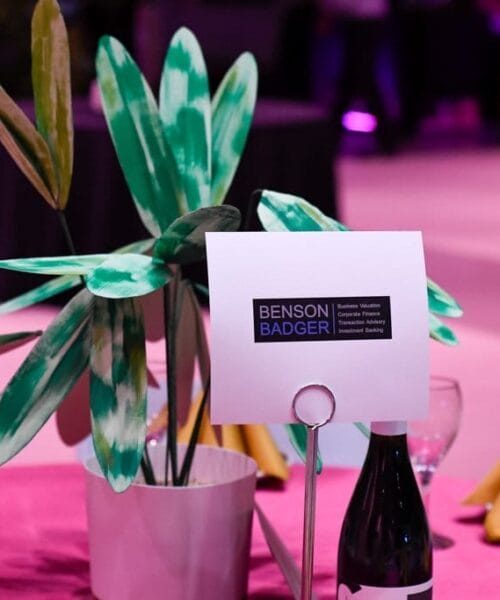 A table set for an event features a teal and green paper flower arrangement, and folded yellow napkins. A card with 