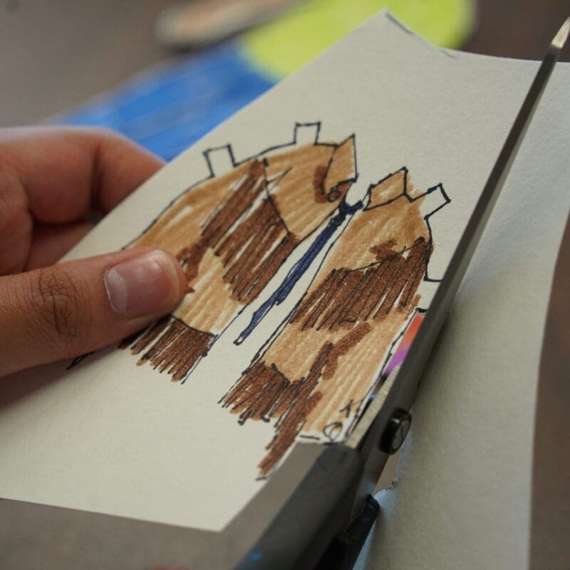A close-up of a hand cutting paper with scissors shows colorful Paper Cutouts of two brown coats or jackets, outlined in black. The blurred background keeps the focus on the vibrant artwork.