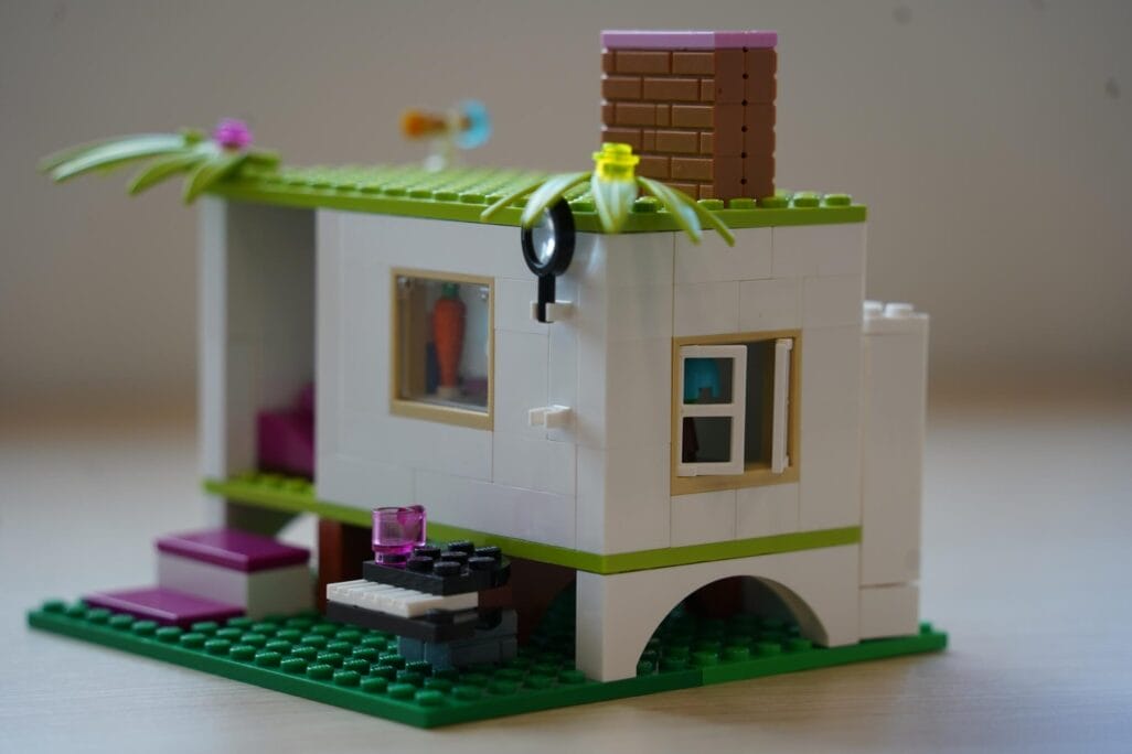 A small LEGO house crafted from building blocks features a white exterior with green and purple accents. It has a window with a figure inside, stairs leading to the entrance, a rooftop with plants, and a chimney. The house also includes a small outdoor table with a pink and black item.