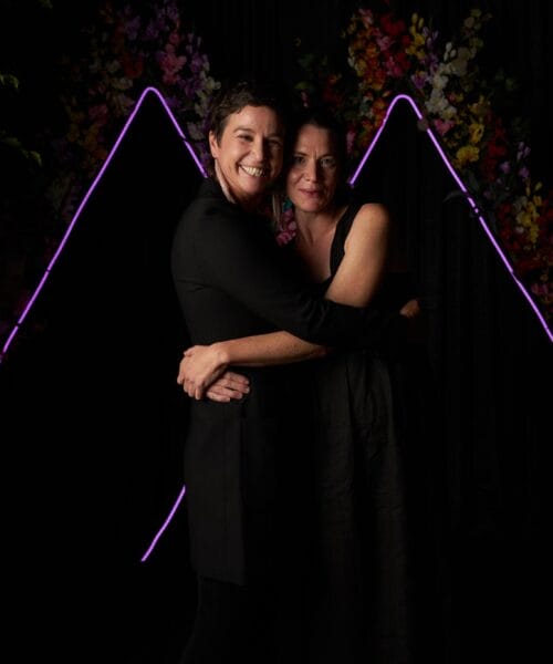 Two people are embracing and smiling warmly at the camera. They are standing in front of a dark backdrop adorned with colorful flowers and glowing purple line art. The overall mood of the photo is joyful and intimate.
