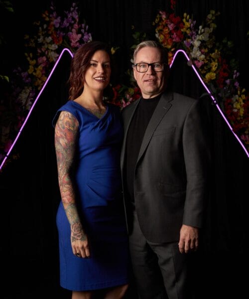 A woman in a blue dress and a man in a black suit and turtleneck stand together in front of a dark backdrop decorated with colorful flowers and pink neon lights. The woman has tattoos on her arms, and they are both smiling at the camera.