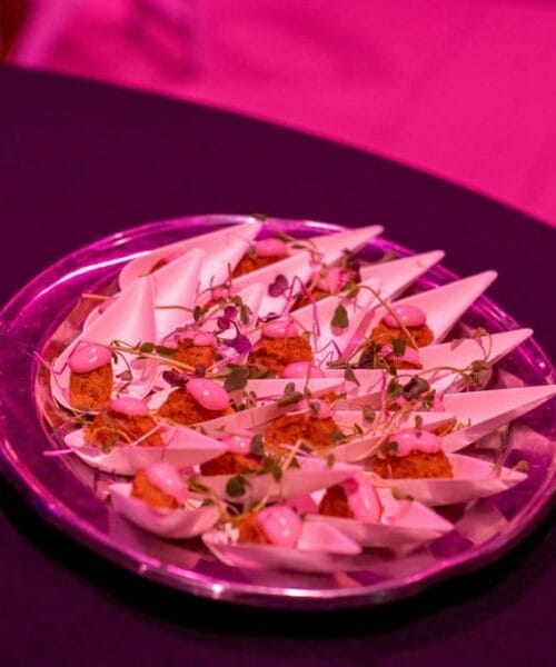A silver platter holds multiple hors d'oeuvres in small, white serving dishes, each featuring a garnish, possibly microgreens, and a dollop of pink sauce. The scene is bathed in a pink light, giving the food and surroundings a vibrant hue.
