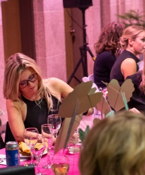 A group of people sit around a table at a social event. Three women in the foreground are engaged in conversation and activities while surrounded by glasses, bottles, and decorations. The background shows additional people and ornate walls, indicating a formal setting.