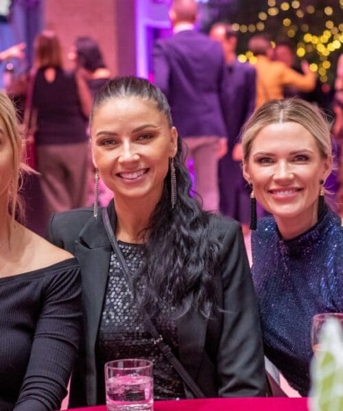 Three women are seated at a table in a lively, festive indoor event. They are smiling and looking at the camera. The background is softly lit, with blurred images of other attendees and decorative lights. A wine bottle and glasses are on the table in the foreground.