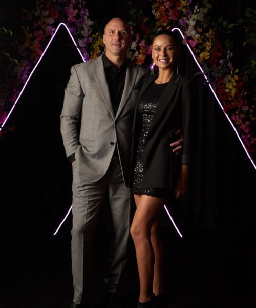 A man and a woman are posing for a photo against a dark backdrop adorned with colorful flowers and illuminated triangle-shaped lights. The man is dressed in a light gray suit with a black shirt, and the woman is wearing a black sequined dress with a black blazer.