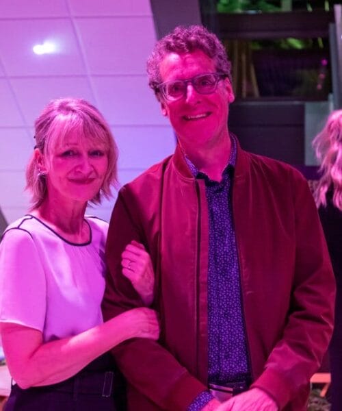 A smiling man and woman stand close together, with the woman's arm around the man's. They are indoors in a modern-looking location with a purple light giving everything a violet tint. Two other people are in the background, engaged in conversation.
