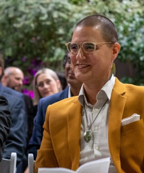 A person with short hair and glasses, wearing a mustard-colored blazer and a white shirt with a necklace, smiles while sitting in an audience. The person next to them is partially visible. The background shows other attendees and some greenery.