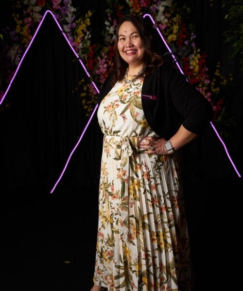 A woman smiling confidently while posing for a photo. She is wearing a floral dress with a belt and a dark cardigan over it. Her outfit is paired with light-colored shoes. The background features neon purple lights and colorful floral decorations.