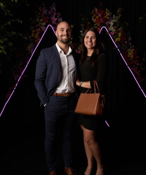 A man in a navy suit and a woman in a black dress stand smiling side by side at a formal event. The woman is holding a brown handbag. The background features a neon triangular outline with colorful floral decorations.