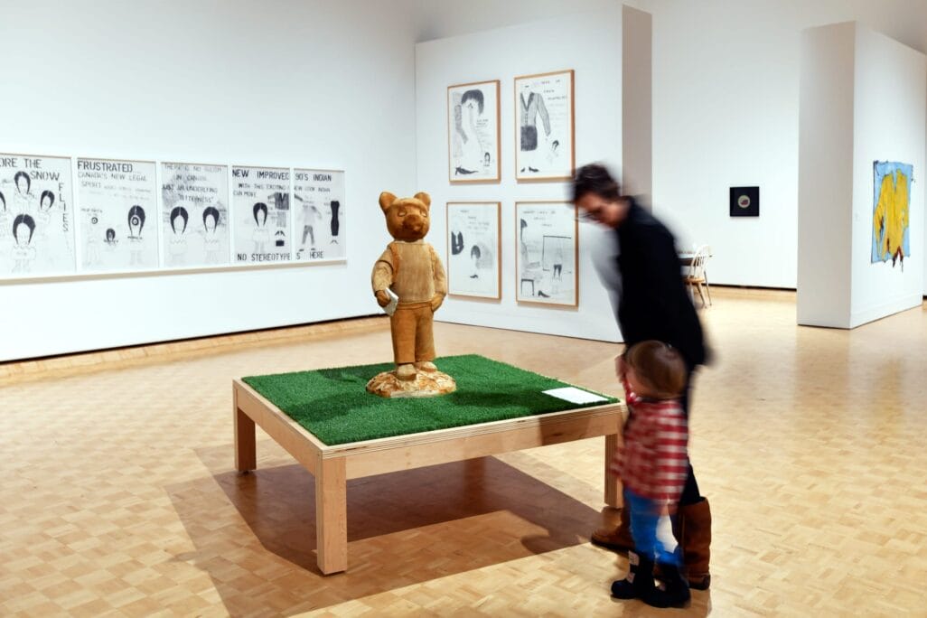 Une personne et un enfant observent une sculpture d'ours sur une plate-forme herbeuse dans une galerie d'art. Diverses œuvres d'art et illustrations encadrées sont exposées sur les murs blancs de la pièce.