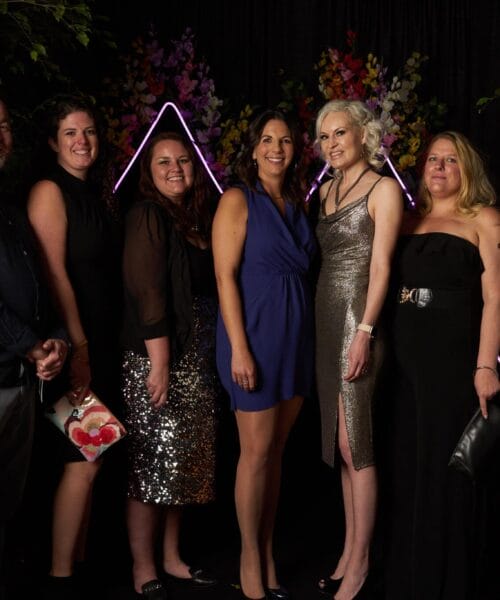 A group of seven people are standing closely together, posing and smiling for the camera. They are dressed in formal attire, with the women wearing elegant dresses and the man in a dark shirt and trousers. The background features neon triangle lights and colorful floral decorations.