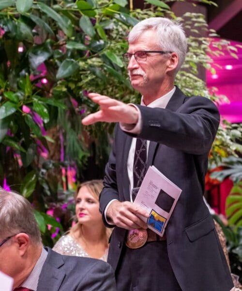 A man wearing glasses and a suit stands with his arm outstretched, holding a piece of paper. He is surrounded by lush greenery and other seated individuals in a well-lit room decorated with pink and purple lights.