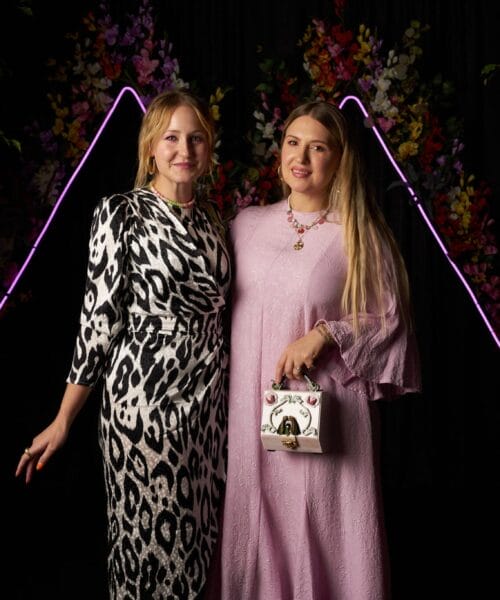 Two women stand closely together, smiling. The woman on the left wears a white and black patterned dress, while the woman on the right wears a lavender dress and holds a small handbag. They are in front of a backdrop with colorful flowers and purple neon lights.