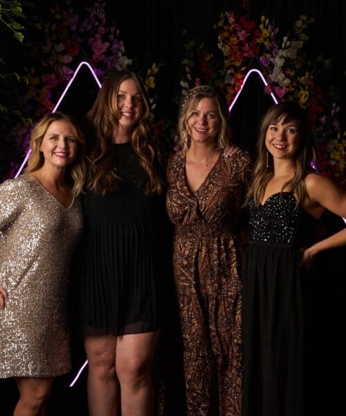 Four women are standing closely together and smiling in front of a dark backdrop adorned with lit geometric shapes and floral arrangements. They are dressed in fashionable evening wear, including a silver sequin dress, a black dress, and patterned outfits.