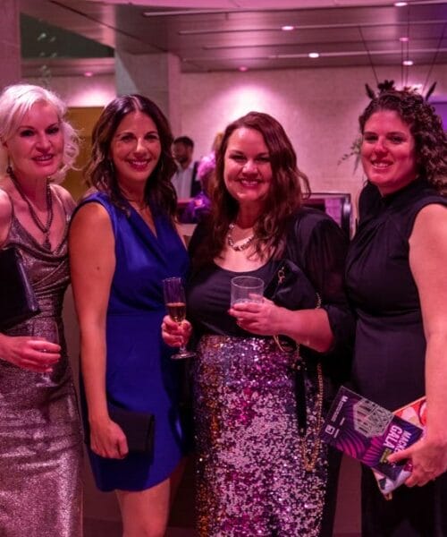 Four women dressed in semi-formal attire are smiling and posing for a photo at an indoor event. The background has a purple-pink lighting, and several other people can be seen socializing. One woman is holding a brochure, and another has a drink in her hand.
