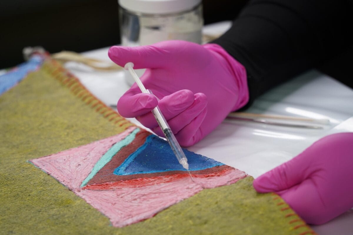 A person in pink gloves uses a syringe to apply a substance to a colorful textile piece with geometric patterns on a table, suggesting restoration or art conservation work.