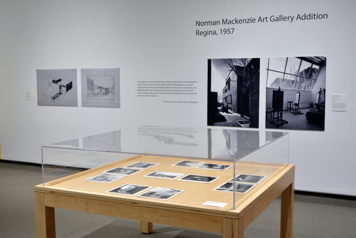 An art gallery display captures the essence of Spring on the Prairie with historical photographs and information about the Norman Mackenzie Art Gallery Addition in Regina, 1957. Images and text adorned on the wall are complemented by additional photos elegantly displayed under glass on a wooden table.
