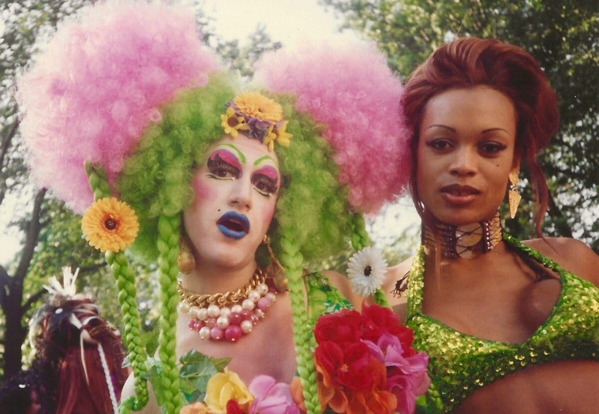 Two individuals participating in a vibrant outdoor event. The person on the left is dressed in a colorful, extravagant outfit with green, pink, and yellow hues, and elaborate makeup. The person on the right is wearing a glittery, green outfit. Trees are visible in the background.