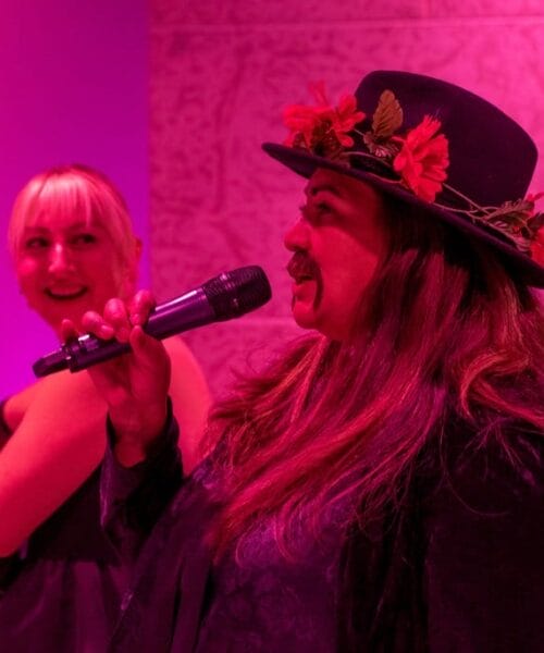 A woman wearing a black hat adorned with flowers speaks into a microphone while another woman in the background looks on and smiles. The image has a pink and purple lighting.