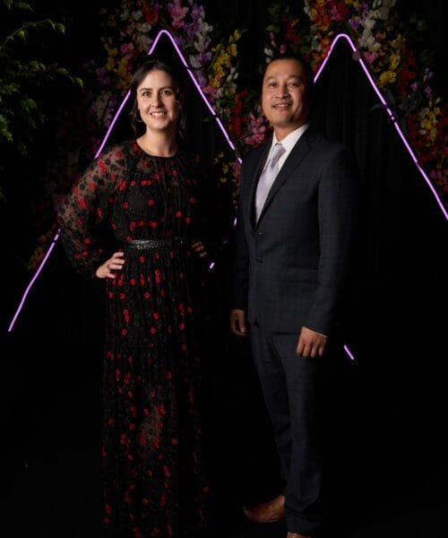 A woman and a man are posing together in a semi-formal setting with colorful floral arrangements and purple neon triangle shapes in the background. The woman is wearing a black floral-patterned dress, and the man is wearing a dark suit with a light-colored tie.