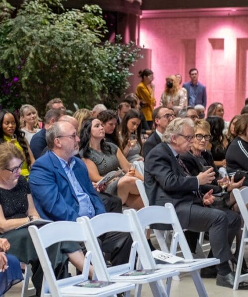 A diverse audience sits on white chairs in an indoor venue, attentively listening. Most are dressed in business or formal attire. The room is decorated with lush greenery and soft lighting. Some attendees hold brochures and drinks. A person stands at the back with a camera.