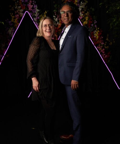 A couple is posing together in front of a dark backdrop with neon triangular lights and vibrant flowers. The woman is wearing a black dress while the man is dressed in a blue suit and a white shirt. They are both smiling and standing close to each other.