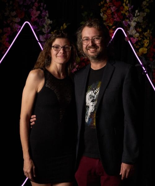 A woman in a black dress and a man in a dark blazer and casual pants stand close together, smiling at the camera. The background features colorful flowers and pink neon triangle lights.