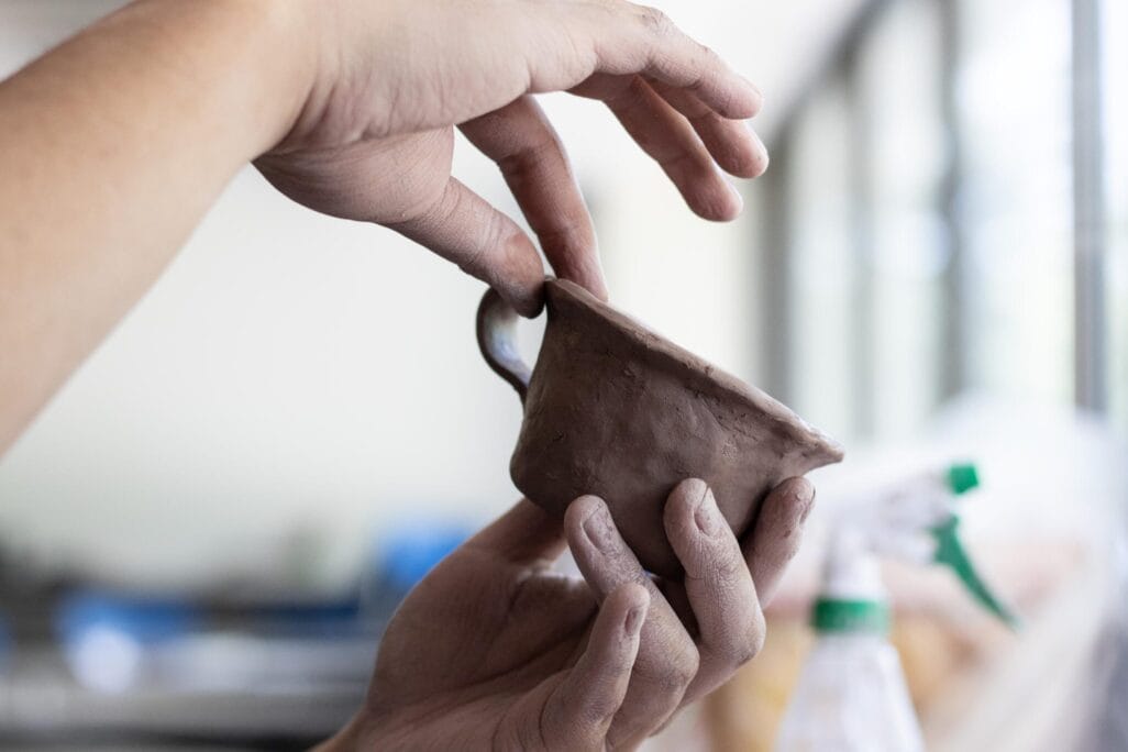 Close-up of hands shaping a small ceramic cup. The fingers are gently smoothing and molding the clay, with a spray bottle visible in the blurred background, suggesting an artistic workspace. This pinch pot portrait captures the precision and craftsmanship involved in pottery.