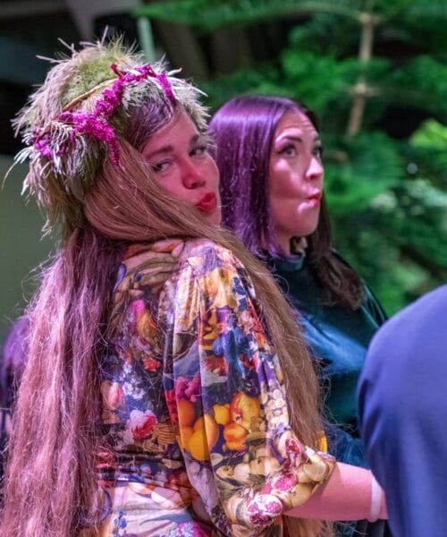 Two women stand in front of a lush, green background. The woman on the left is wearing a floral dress and a natural headpiece, with long hair cascading over her shoulders. The woman on the right has straight, dark hair and is dressed in dark clothing. Both women are looking sideways.