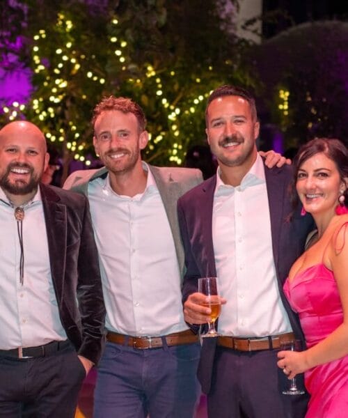 A group of five people is smiling and posing together at an upscale, brightly lit outdoor event. They are dressed in formal attire, with two women in dresses and three men in suits. The background features trees with sparkling lights and a purple ambiance.