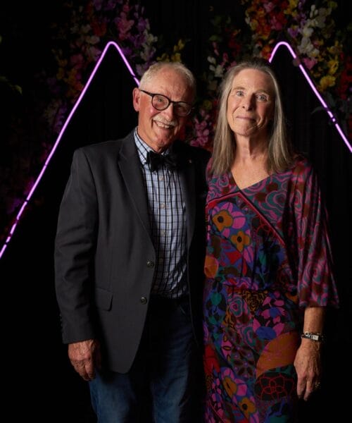 An older man and woman stand together, smiling in front of a dark backdrop with colorful flowers and purple neon triangle lights. The man is wearing a dark blazer and bow tie, while the woman is in a colorful patterned dress. Trees are visible on either side of them.