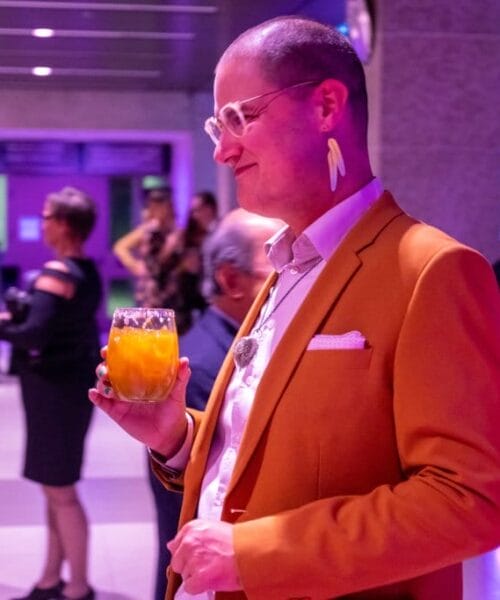 A bald man in a mustard-colored blazer, white shirt, and statement earrings is holding a drink in a glass filled with an orange liquid. He stands at a lively indoor event, with other people socializing in the background under purple lighting.