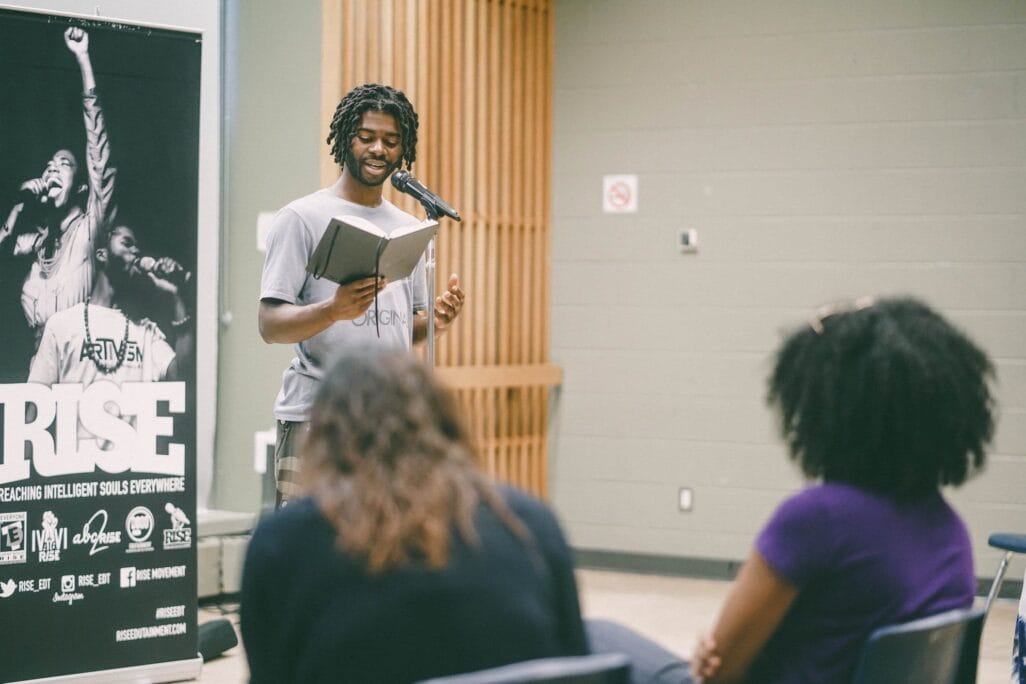 A person with locs reads from a notebook at a microphone in front of an audience. Behind them is a large banner displaying the word 
