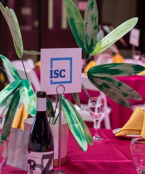 A round banquet table covered with a pink tablecloth, set with yellow napkins, wine glasses, and a bottle of wine. A centerpiece features green leaves and a sign with 