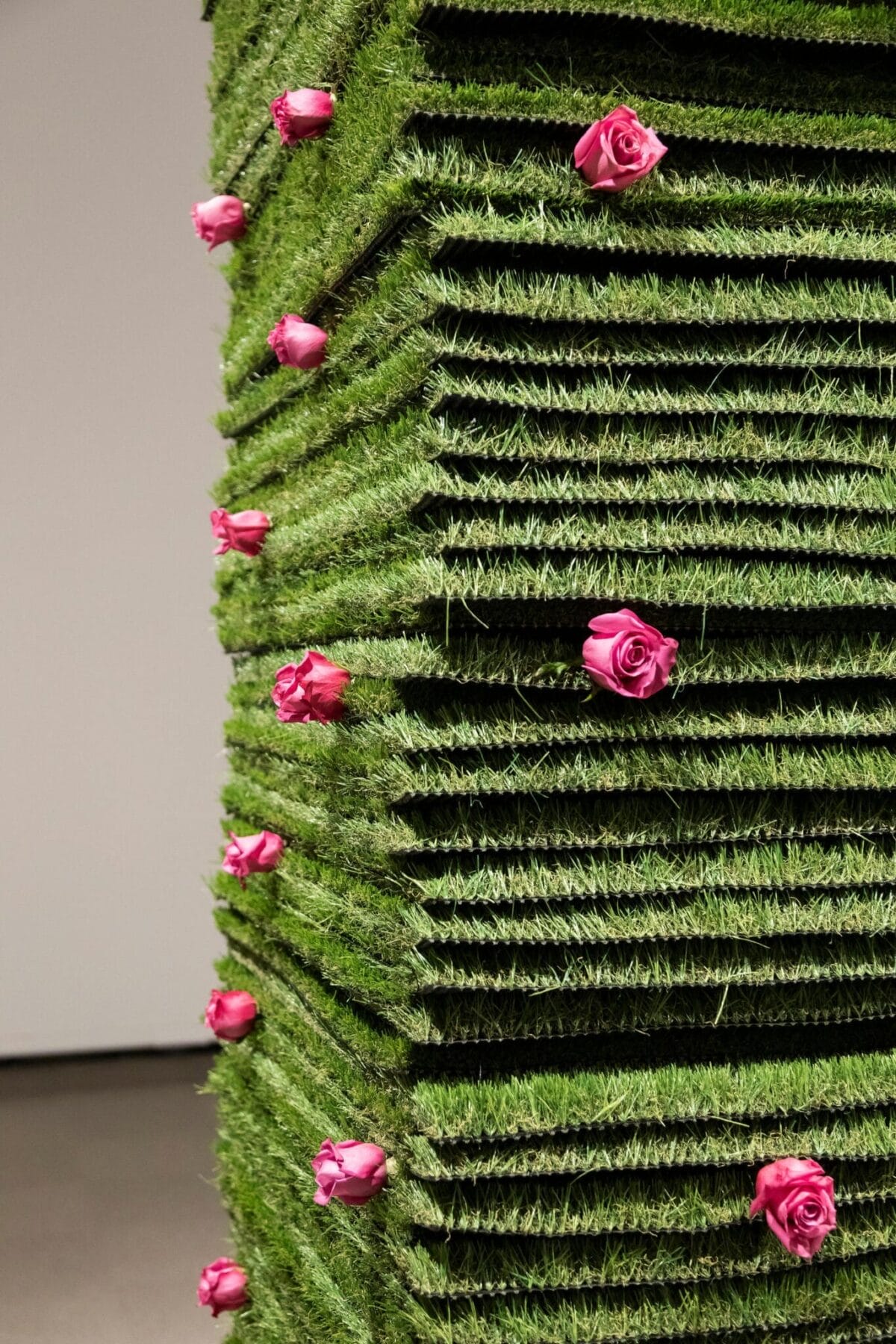 A vertical stack of square, green grass mats adorned with several pink roses embedded in various layers. The background is a plain, light-colored wall. The arrangement creates a visually striking contrast between the vibrant green grass and the pink roses.