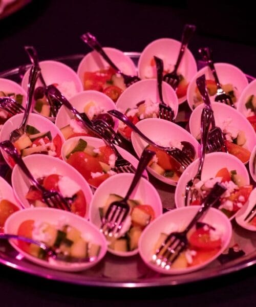A platter of small white bowls, each containing a fresh salad with diced tomatoes, cucumbers, and cheese, arranged neatly. Every bowl has a small fork, ready for individual servings. The scene is illuminated with pink lighting, suggesting a festive environment.