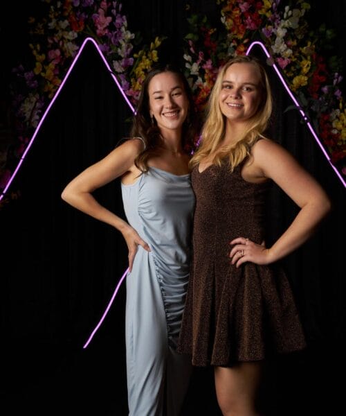 Two women pose smiling in elegant dresses against a black backdrop with purple accent lighting shaped like abstract mountains. One woman wears a light blue dress with a side slit, and the other wears a dark brown dress. Green foliage and flowers decorate the background.