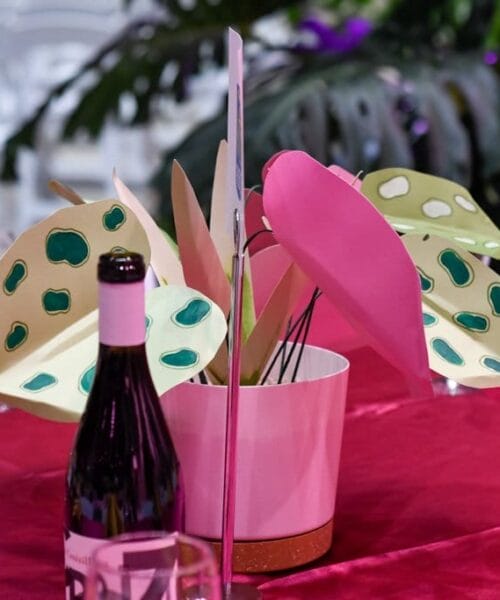 A table setting with a red tablecloth features a central white pot with colorful paper leaves, some pink and some with green spots. Surrounding the pot are wine glasses, orange folded napkins, and a bottle of red wine. In the background, greenery is visible.