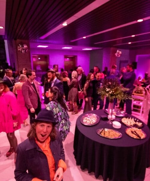 A lively indoor party with numerous people socializing under vibrant purple lighting. Guests are dressed in colorful attire, chatting and mingling around a table filled with various appetizers. The atmosphere appears joyful and celebratory.