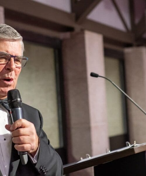 An older man in a tuxedo speaks into a microphone while standing at a podium. He wears glasses and has short, gray hair. In the background, a large screen displays content, and the setting appears to be a formal event or conference.