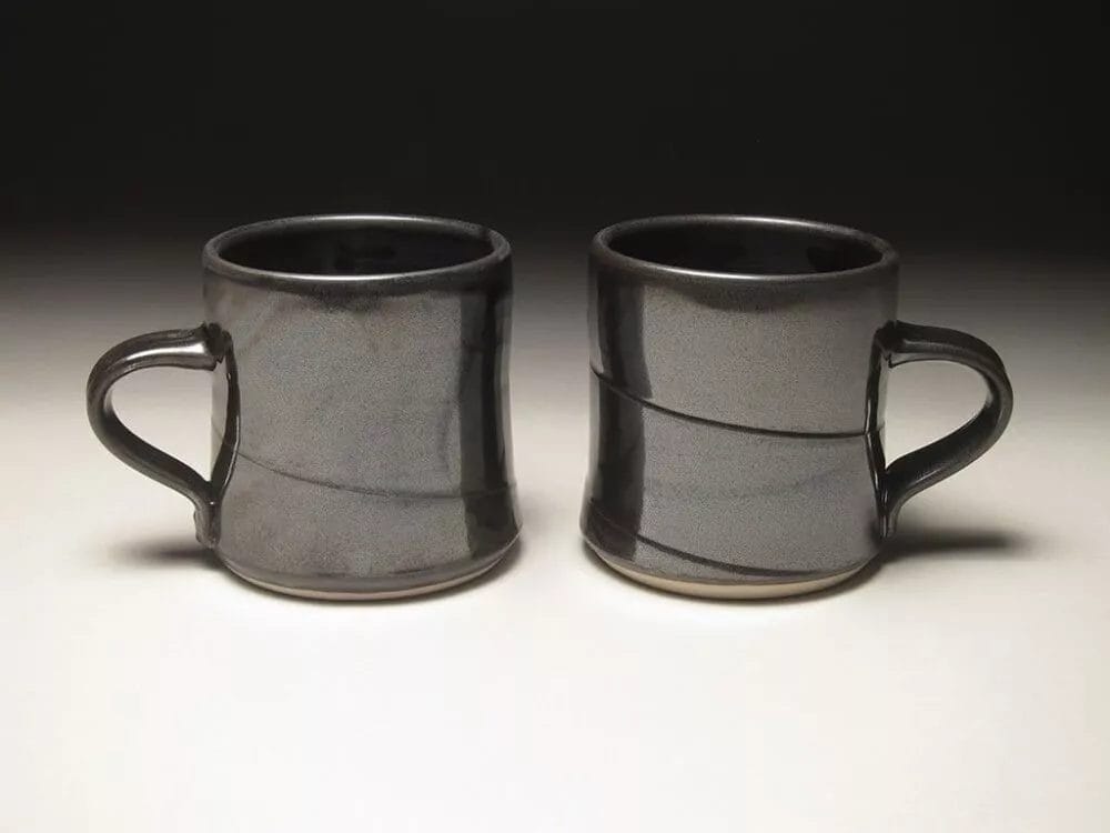 Two dark gray *Mugs by Zane Wilcox* with handles are placed side by side on a neutral surface. Each mug has a slightly flared base and a subtle horizontal groove around the middle. The background is a gradient from light to dark, highlighting the mugs.