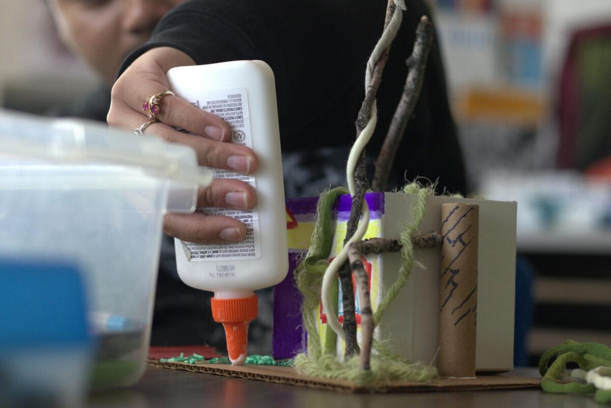A person is using a bottle of white glue to assemble a creative pinch pot project. The activity involves sticks, yarn, a cardboard tube, and other materials, suggesting an engaging scene in a classroom or craft area.