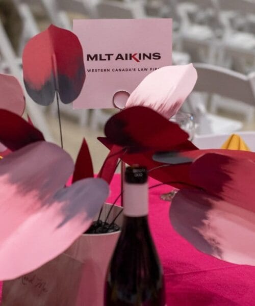 A table centerpiece featuring red and pink painted leaves is positioned on a pink tablecloth. A bottle of wine is in the foreground. At the top, there is a card with the text 